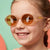 Happy and confident little girl with freckles wearing SmileyWorld acorn brown oval kids sunglasses with yellow smiley face details on the temples, against a split light green and peach background.