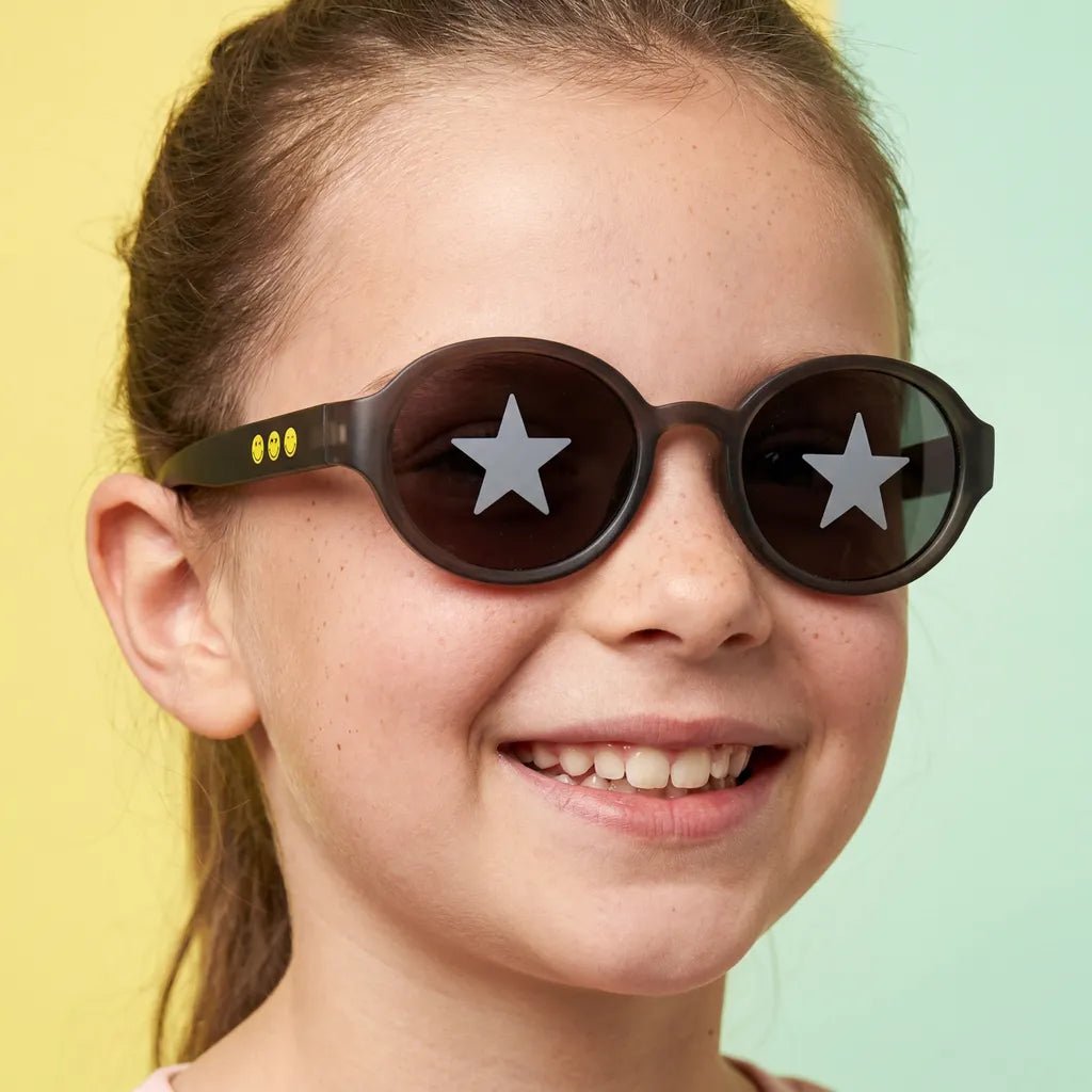 Happy and smiling little girl with freckles wearing SmileyWorld obsidian black oval kids sunglasses with yellow smiley face details on the temples and star designs on the lenses, against a split bright yellow and light green background.