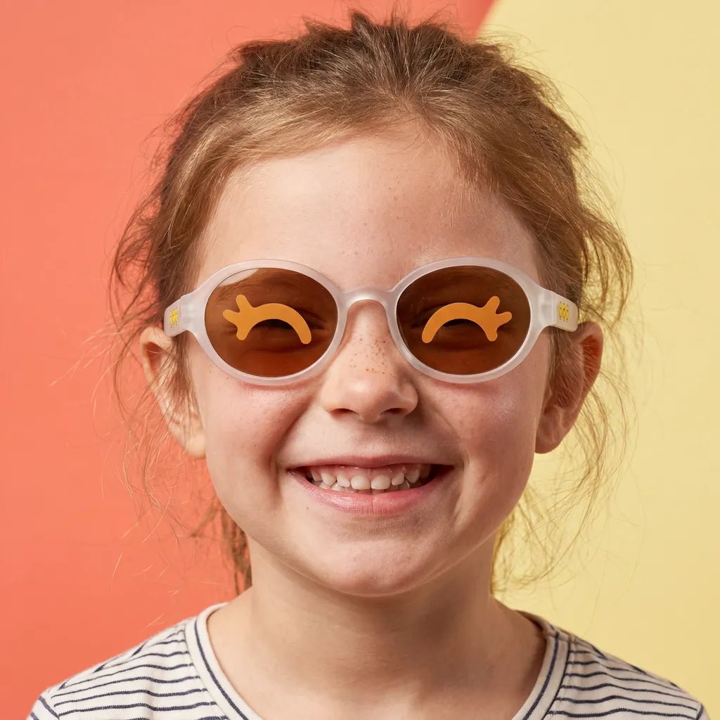 Happy and smiling little girl with freckles wearing SmileyWorld snow white oval kids sunglasses with yellow smiley face details on the temples and closed happy eyes designs on the lenses, dressed in a striped shirt against a split orange and yellow background.
