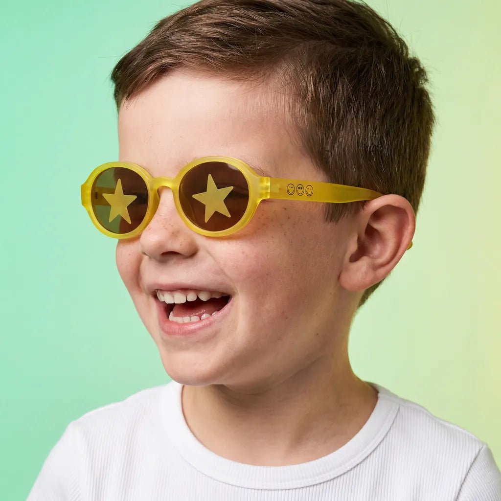 Happy and laughing little boy with freckles wearing SmileyWorld sunlight yellow oval kids sunglasses with yellow smiley face details on the temples and star designs on the lenses, dressed in a white t-shirt against a light green and yellow gradient background.