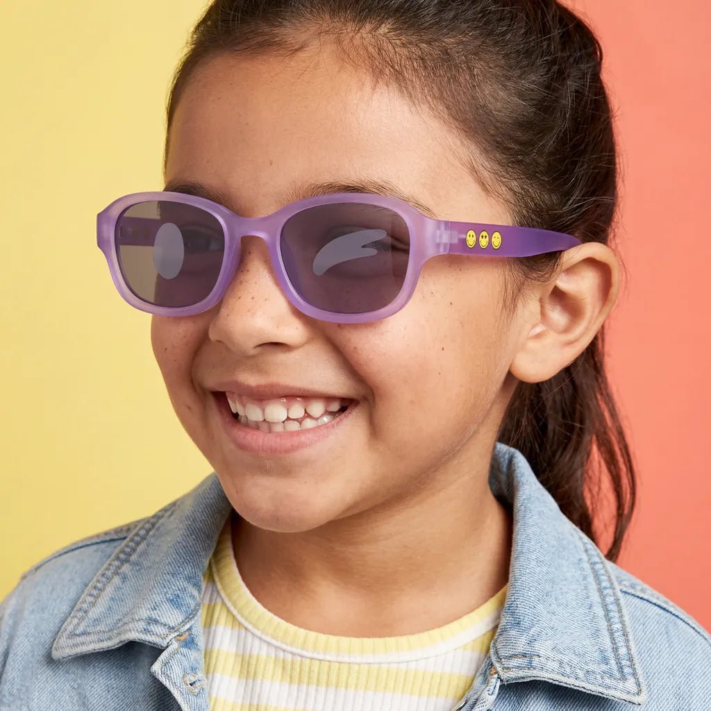Happy and confident little girl with freckles wearing SmileyWorld coral purple square kids sunglasses with yellow smiley face details on the temples, dressed in a denim jacket against a bright yellow and orange background.