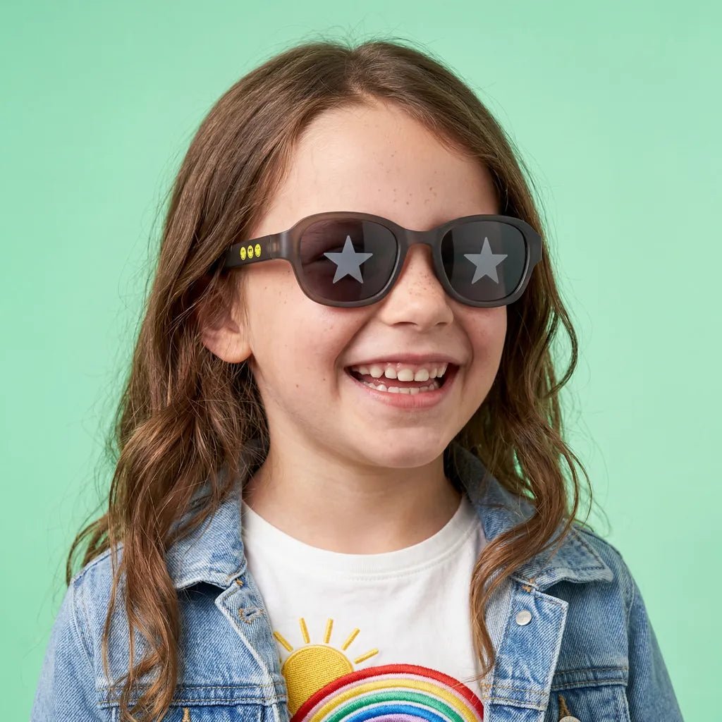 Happy and laughing little girl with freckles wearing SmileyWorld obsidian black square kids sunglasses with yellow smiley face details on the temples and star designs on the lenses, dressed in a white t-shirt with a rainbow graphic and a denim jacket against a solid light green background.