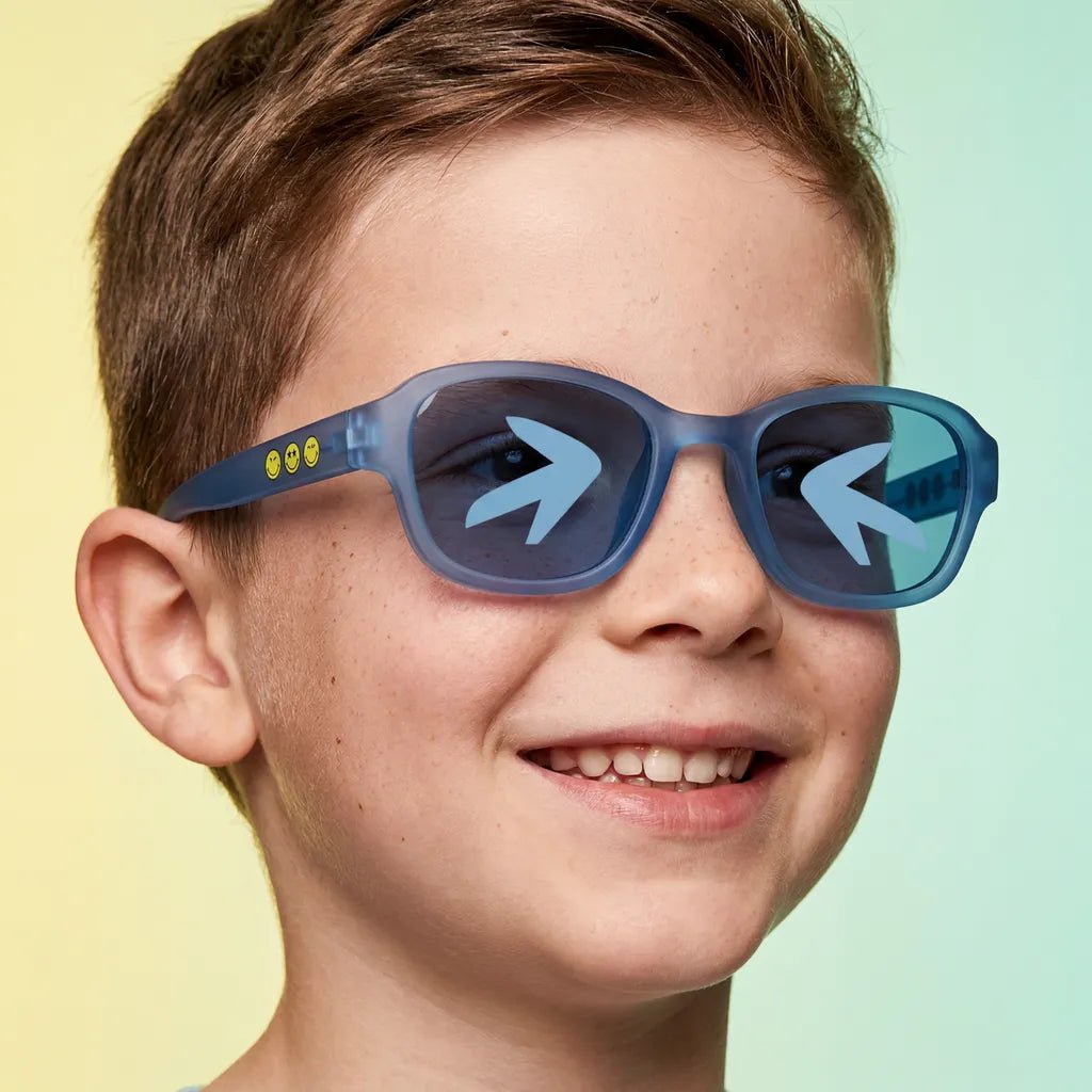 Happy and smiling little boy with freckles wearing SmileyWorld sea blue square kids sunglasses with yellow smiley face details on the temples and arrow designs on the lenses, against a yellow and light green gradient background.