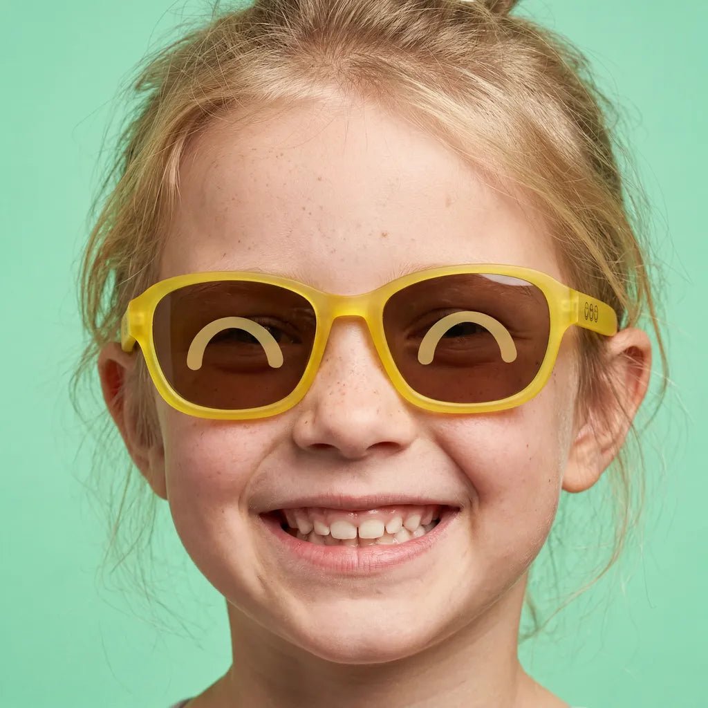 Happy and smiling little girl with freckles wearing SmileyWorld sunlight yellow square kids sunglasses with yellow smiley face details on the temples and closed happy eyes designs on the lenses, against a solid light green background.